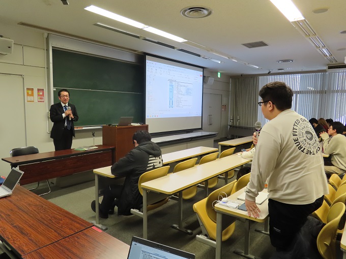 教室で講師が前方で説明し、学生が席から立って質問している様子