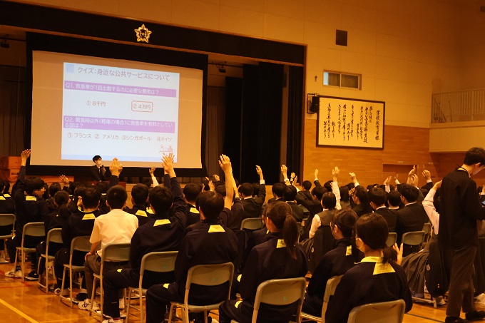 スクリーンに映った資料を見ながら手を挙げてクイズに答える生徒達（平岡緑中学校）
