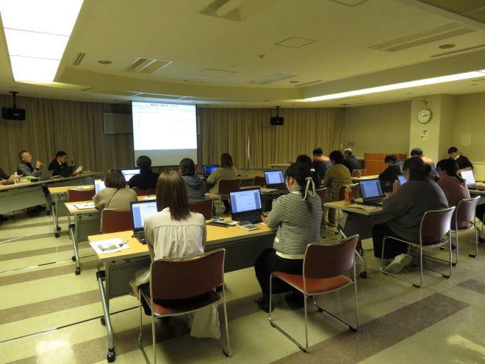 会場で複数の参加者が資料を見ながら話しを聞いている様子