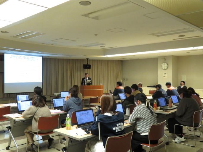 会場で前方に登壇者、後方に参加者が並び、発表を聞いている様子