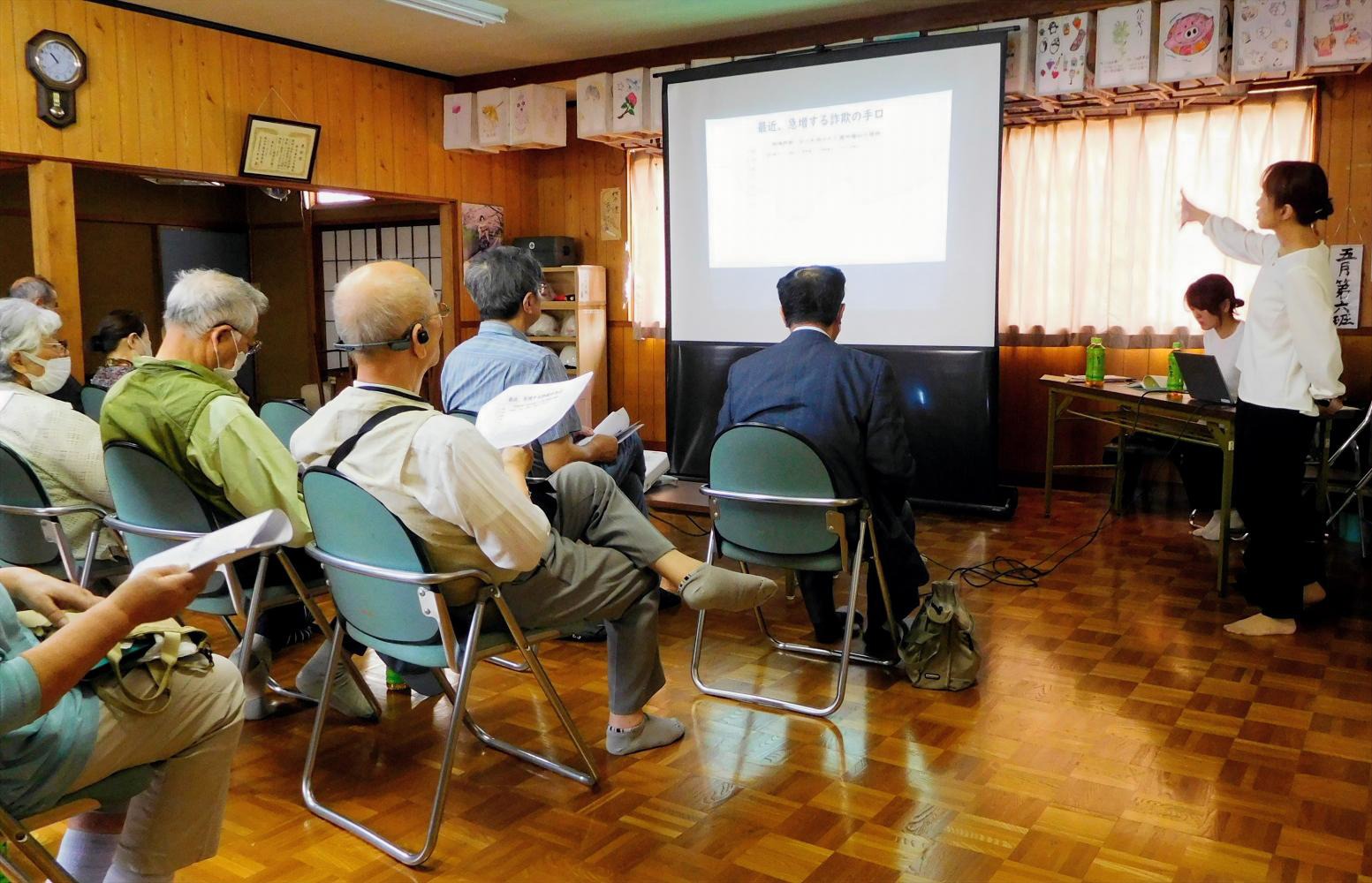 財務事務所の女性職員が高齢者の前方に立ってスクリーンに投影した資料をもとに特殊詐欺等の注意喚起を行っている様子