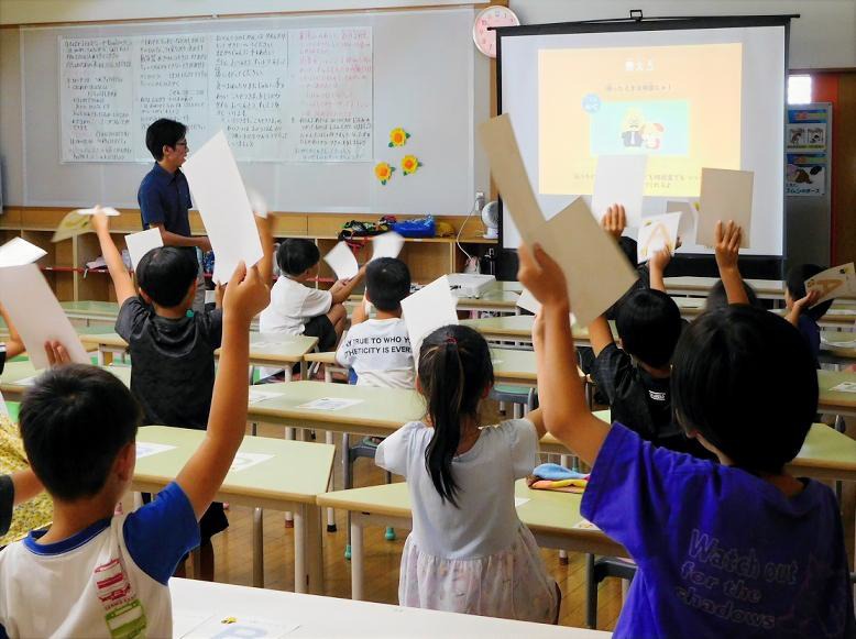 うんこお金ドリルクイズ大会に参加している児童がクイズの正解だと思うカードを上げている様子