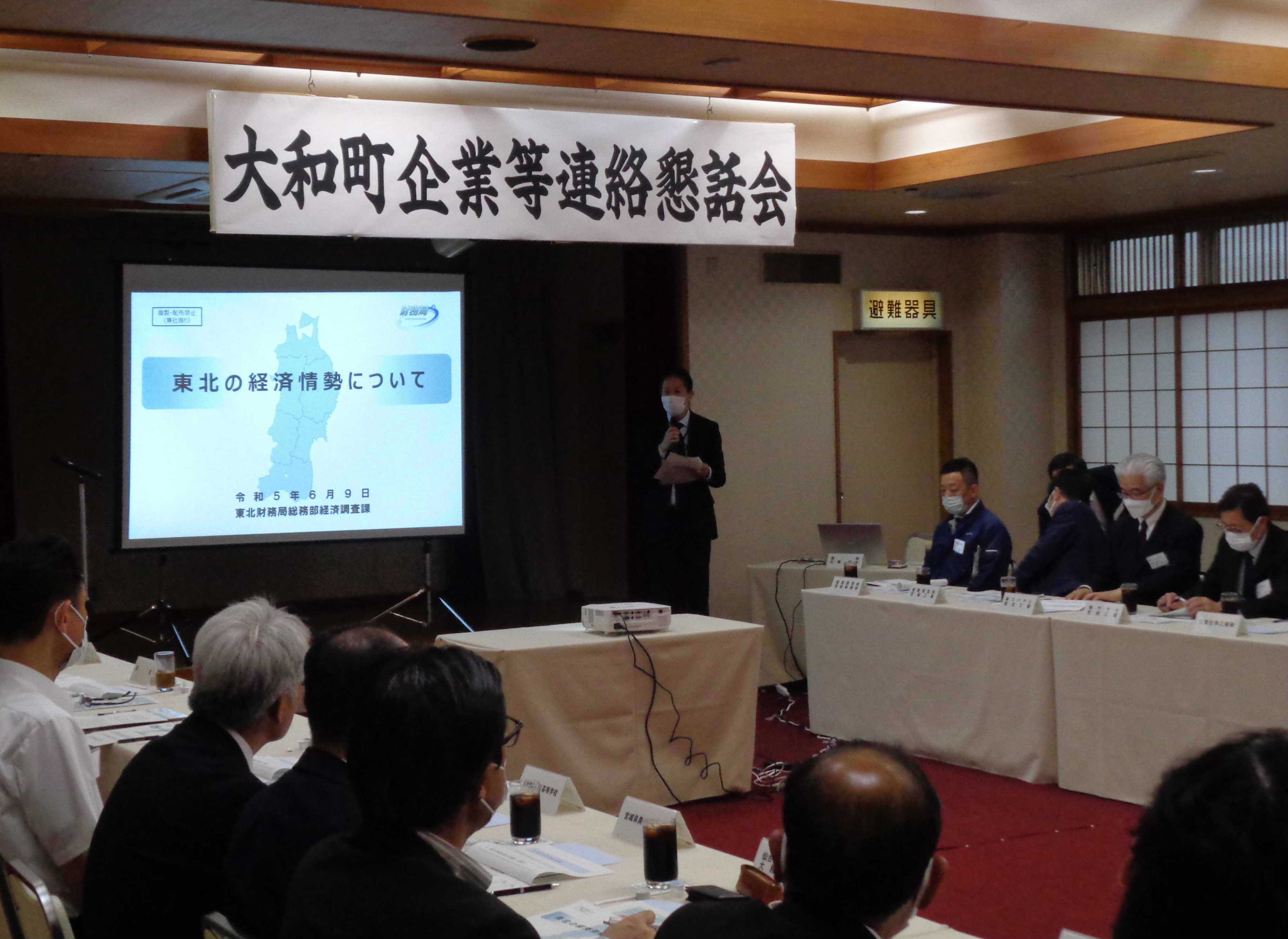 男性職員が旅館の和室にて、大和町の企業者数十人の前で講演している様子