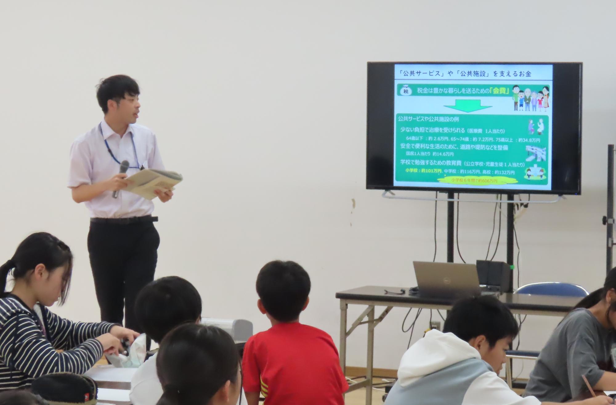 財務局職員がスクリーンを使用して児童に講義をしている様子