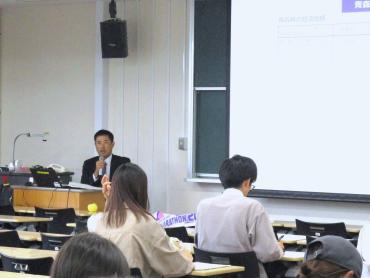 前方にスクリーンと黒板のある教室で、講師が受講者に向けて説明している様子