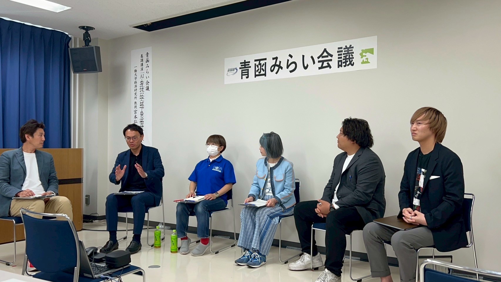 パネリスト5人が着席し、左端の眼鏡の男性が意見を述べる様子