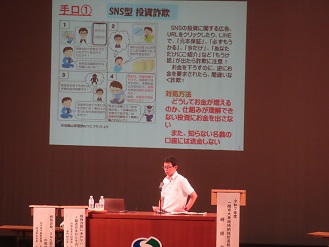 SNS型投資詐欺のスライドを投影し、壇上で説明する写真