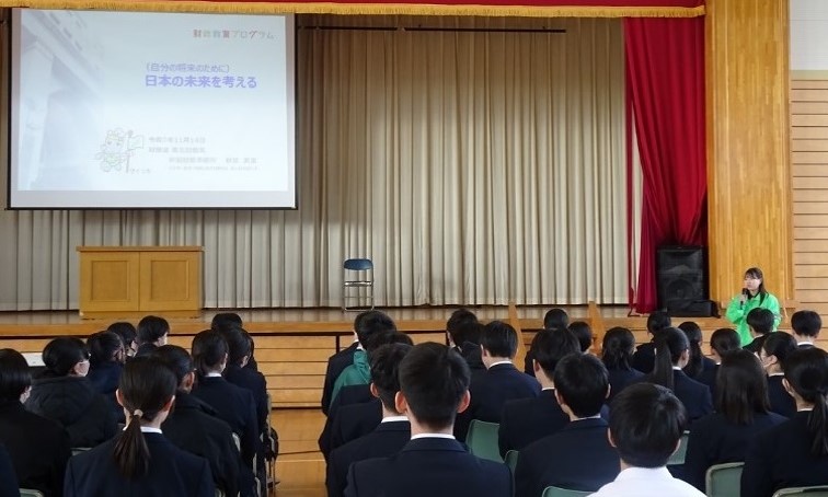 財務事務所職員が体育館のスクリーンに投影したスライドを使用し財政の説明を行っている様子