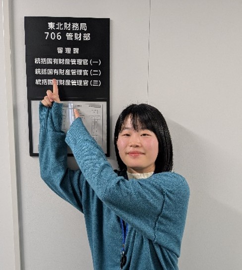 廊下の管財部の看板を笑顔で指さす女性職員