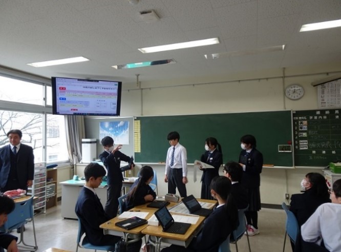 生徒がグループで作成した予算案を黒板の前で発表している様子