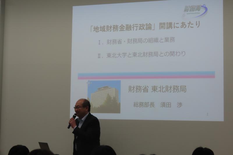 財務局の講師が、大勢の学生に向けてスクリーンを使用して講義をしている様子