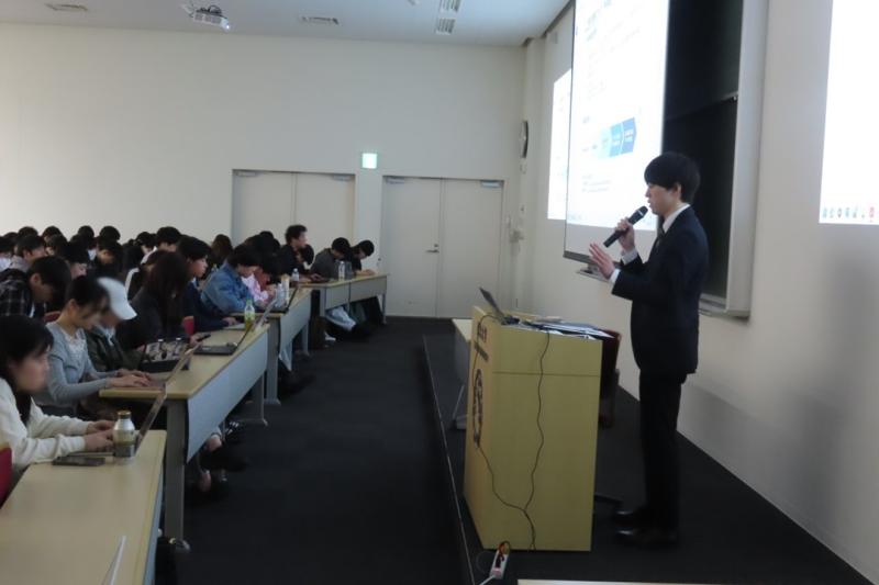 東北大学OBの講師が、スクリーンを使用し、マイクを持って大勢の学生の前で講義をする様子