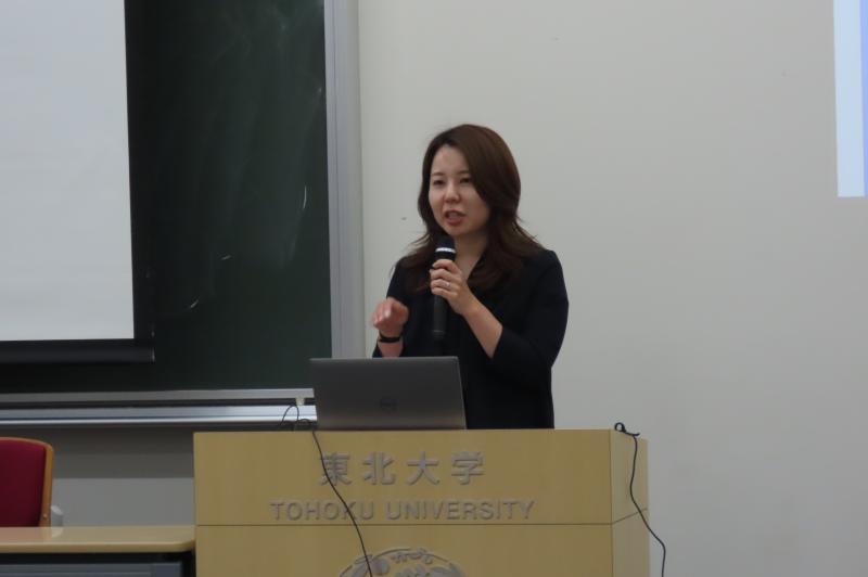 財務局の講師が、壇上でマイクを持って講義をする様子