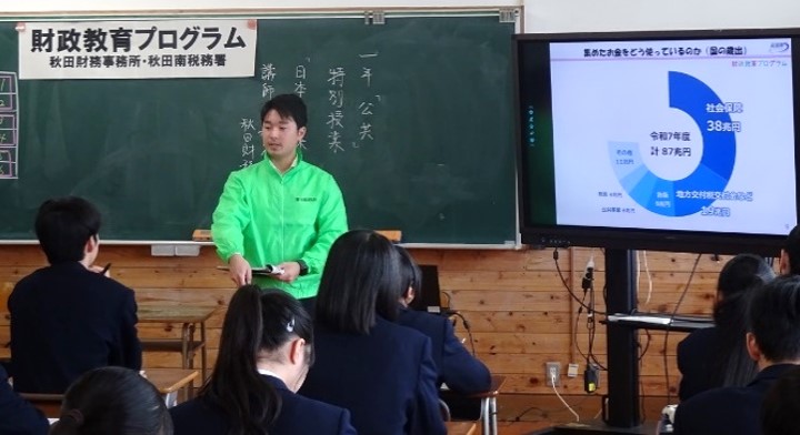 財務事務所職員が教室のモニターに投影した円グラフで財政の説明を行っている様子