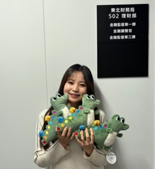 廊下の理財部の看板前にワニーサのぬいぐるみを３つ持って笑顔を見せる女性職員